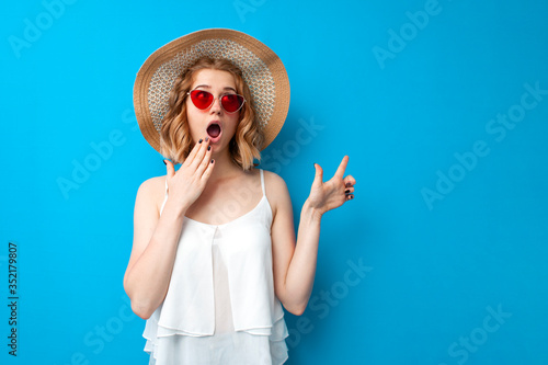 summer concept. shocked girl in a sun hat and glasses shows a copy space on a blue isolated background, emotional woman in summer clothes on vacation