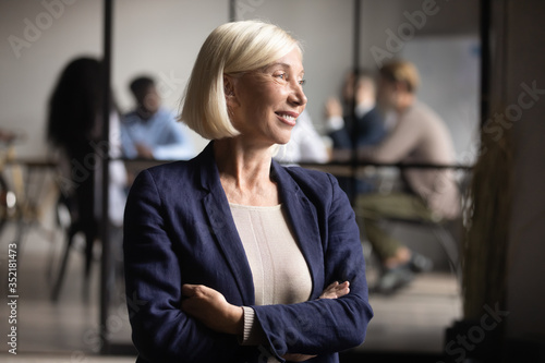 Head shot smiling middle aged businesswoman dreaming about good future, pondering new project strategy, looking in distance, standing with arms crossed in modern office, business vision concept