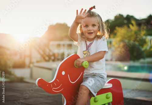 Happy baby child riding a red elephant at the playground and waving his hand.
