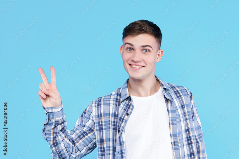 Cheerful smiling guy dressed in white t-shirt, checkered shirt on blue background. Young man is showing with fingers number two, peace sign, discount. Male gesturing. Emotional portrait concept. Cheerful smiling guy dressed in white t-shirt, checkered shirt on blue background. Young man is showing with fingers number two, peace sign, discount. Male gesturing. Emotional portrait concept.