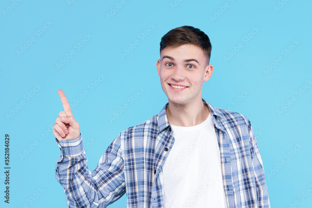 Cheerful smiling guy dressed in white t-shirt, checkered shirt on blue background. Young man is showing with fingers number one, percents, discount. Male gesturing. Emotional portrait concept. Cheerful smiling guy dressed in white t-shirt, checkered shirt on blue background. Young man is showing with fingers number one, percents, discount. Male gesturing. Emotional portrait concept.