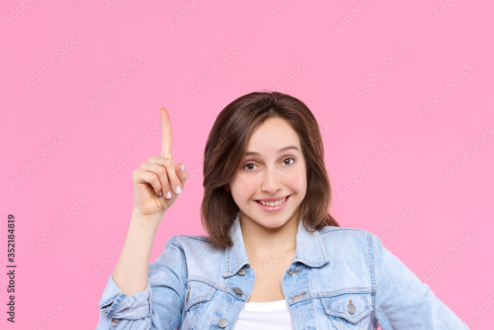 Cheerful smiling girl dressed in white t-shirt, denim jacket is standing on pink background. Young woman is showing direction with hand to up. Female gesturing. Emotional portrait concept. Cheerful smiling girl dressed in white t-shirt, denim jacket is standing on pink background. Young woman is showing direction with hand to up. Female gesturing. Emotional portrait concept.