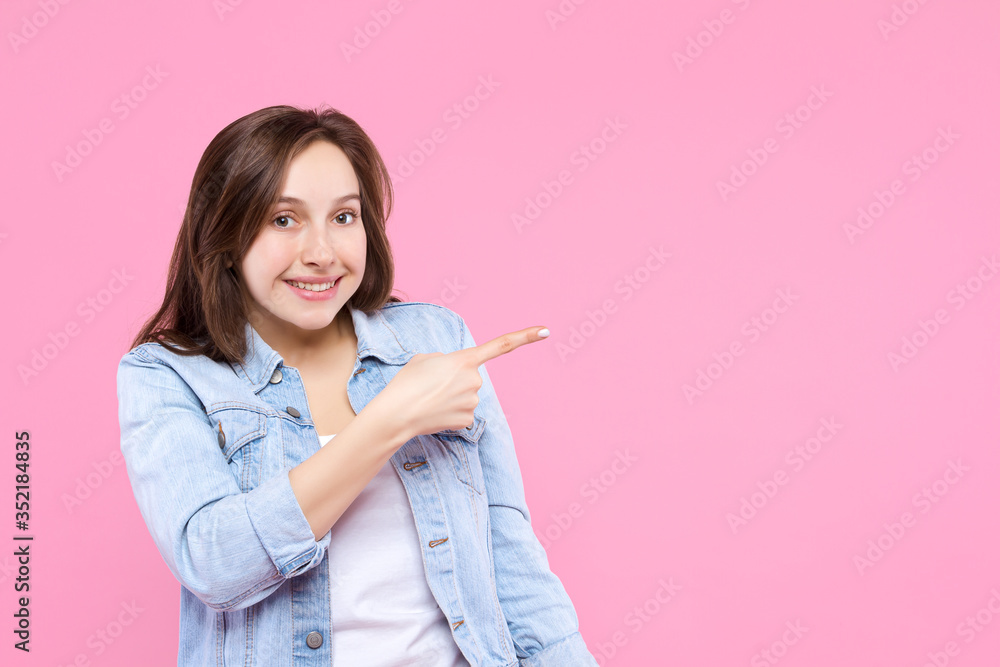 Pretty smiling girl dressed in white t-shirt, checkered denim jacket is standing on pink background. Young woman is showing direction with hand to right. Female gesturing. Emotional portrait concept. Pretty smiling girl dressed in white t-shirt, checkered denim jacket is standing on pink background. Young woman is showing direction with hand to right. Female gesturing. Emotional portrait concept.