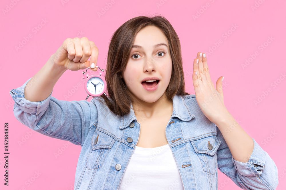 Pretty puzzled surprised girl in white t-shirt, denim jacket is holding small alarm clock. Young woman is showing what time, standing on pink background. Emotional portrait. Menstrual cycle concept. Pretty puzzled surprised girl in white t-shirt, denim jacket is holding small alarm clock. Young woman is showing what time, standing on pink background. Emotional portrait. Menstrual cycle concept.