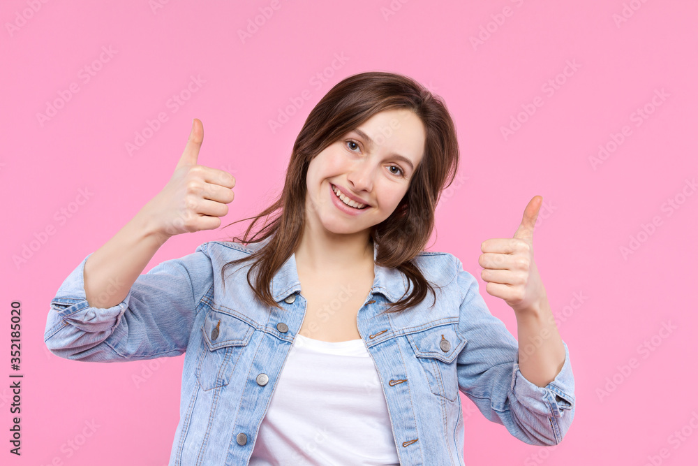 Satisfied pleased girl dressed in white t-shirt, denim jacket is standing on pink background. Young woman is showing positive thumbs up sign. Female gesturing. Emotional portrait concept. Satisfied pleased girl dressed in white t-shirt, denim jacket is standing on pink background. Young woman is showing positive thumbs up sign. Female gesturing. Emotional portrait concept.