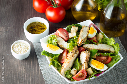Fresh Caesar salad made of tomato, ruccola, chicken breast, eggs, arugula, crackers and spices. Organic ingredients in a white, transparent bowl on wooden background