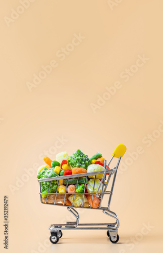 Frozen vegetables assorted in toy shopping cart on cream background. Full of assorted frozen vegetables food shop trolley at beige or yellow backdrop. Minimalistic concept. Vertical. Copy space