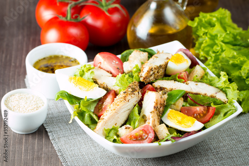 Fresh Caesar salad made of tomato, ruccola, chicken breast, eggs, arugula, crackers and spices. Organic ingredients in a white, transparent bowl on wooden background