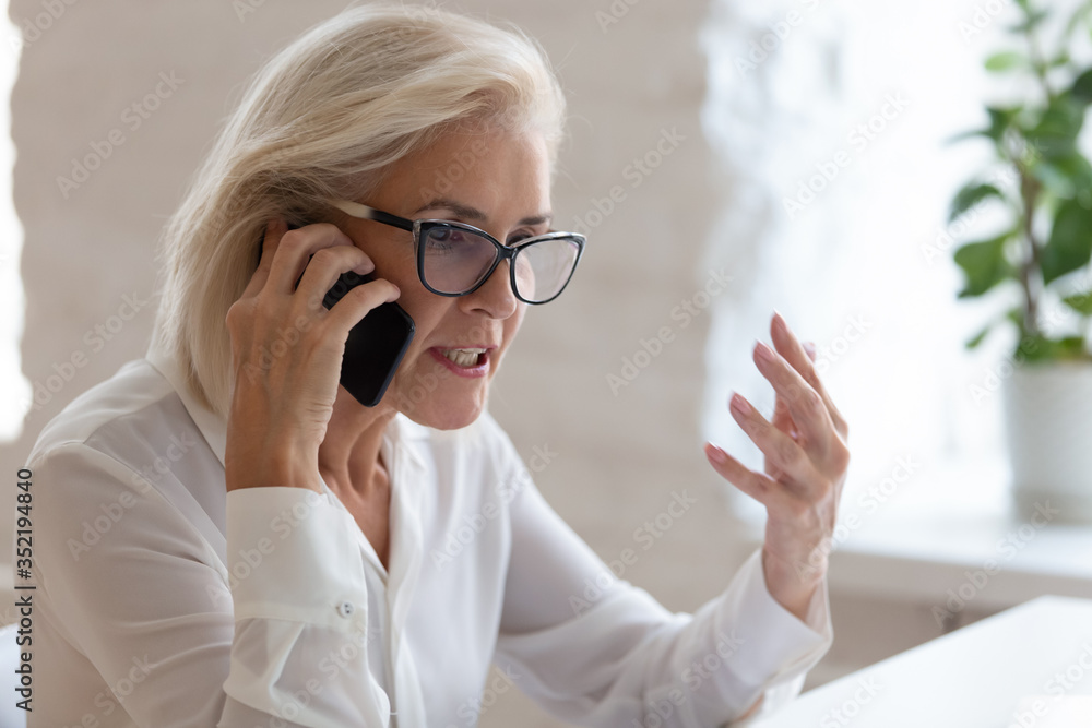 Angry senior female employee in glasses argue with client or customer talking on smartphone, mad frustrated middle-aged businesswoman feel distressed dispute solve problem over cellphone call