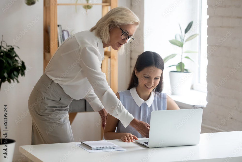 Successful middle-aged businesswoman help supervise young intern work on laptop, confident senior lady boss explain computer job project to millennial female employee at workplace, teamwork concept Successful middle-aged businesswoman help supervise young intern work on laptop, confident senior lady boss explain computer job project to millennial female employee at workplace, teamwork concept