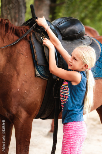 Fototapeta Naklejka Na Ścianę i Meble -  Cute young girl riding a brown horse on a country road at sunset. The child quietly travels on a stallion. The girl confidently controls the horse in a blooming field. Children's summer sports camp.