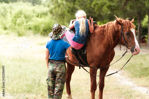 The instructor helps the girl to saddle a brown horse in the forest. Children's equestrian camp. Summer sports camp for children. A nice girl is learning to ride a horse. © anantaradhika