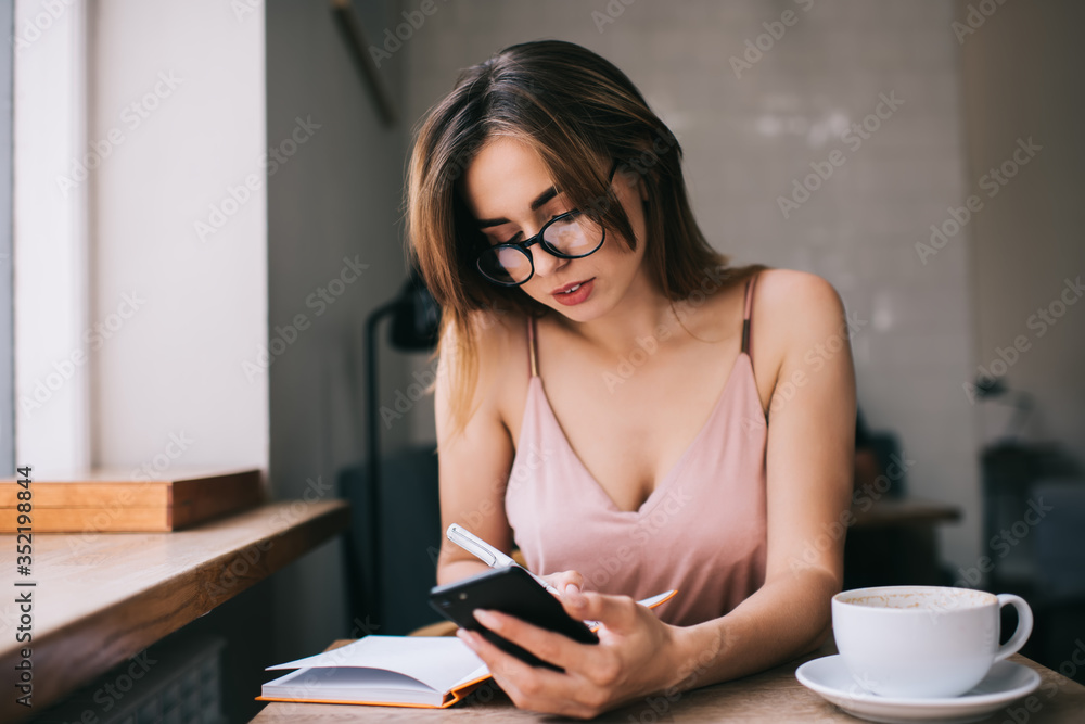 Focused young woman taking notes in daily planner while using smartphone in cafe Focused young woman taking notes in daily planner while using smartphone in cafe