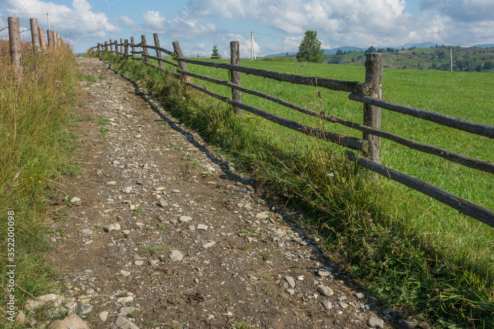 A country road leads up between the wooden fences. In the distance, mountains and a village are visible.