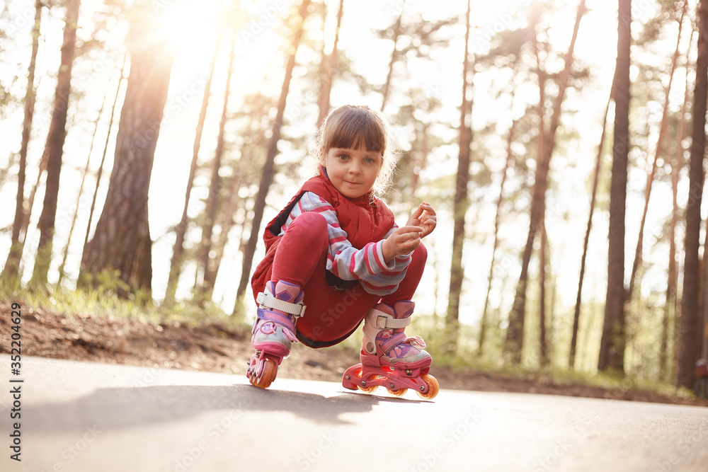 Picture of funny pleasant pretty kid sitting on road in inline skates, looking directly at camera, spending time alone, having pensive facial expression, wearing casual clothes. Childhood concept. Picture of funny pleasant pretty kid sitting on road in inline skates, looking directly at camera, spending time alone, having pensive facial expression, wearing casual clothes. Childhood concept.