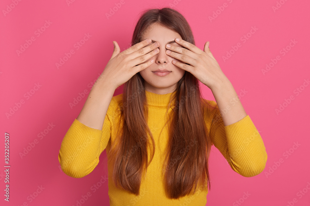 Horizontal shot of peaceful good looking cute young model standing isolated over pink background in studio, covering eyes with hands, wearing yellow sweatshirt, being alone. Surprise concept. Horizontal shot of peaceful good looking cute young model standing isolated over pink background in studio, covering eyes with hands, wearing yellow sweatshirt, being alone. Surprise concept.