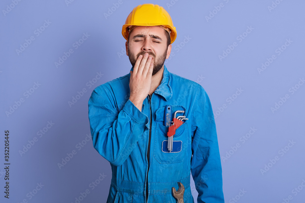Image of sleepy exhausted builder yawning, covering his mouth with hand, closing eyes, having beard, wearing uniform and helmet, being tired of work, having desire to sleep. People and work concept. Image of sleepy exhausted builder yawning, covering his mouth with hand, closing eyes, having beard, wearing uniform and helmet, being tired of work, having desire to sleep. People and work concept.