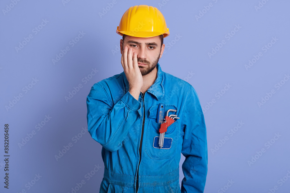 Portrait of upset disappointed sad young builder having frowned facial expression, losing job on account of coronavirus outbreak, putting hand on cheeck, wearing blue uniform and yellow helmet.