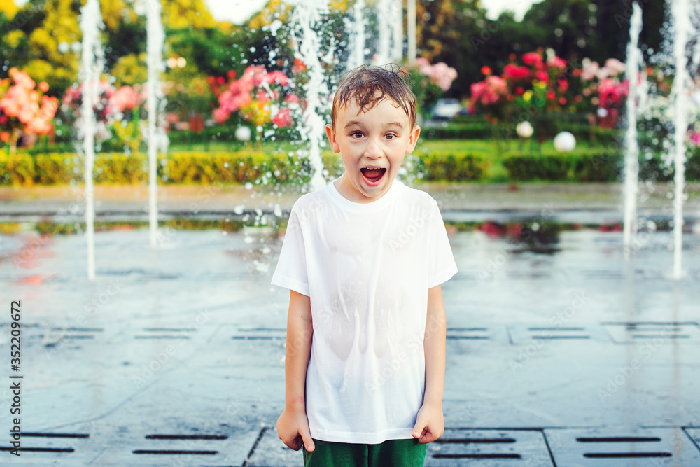 Naklejka premium Happy boy plays in the square between the water jets in the fountain at synny summer day. Summer vacation, lifestyle.