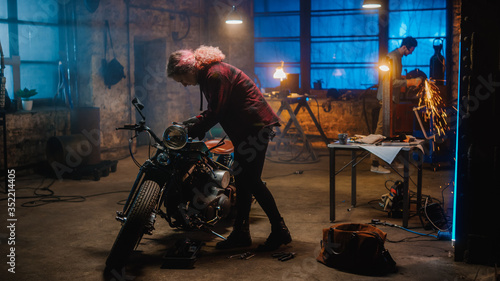 Young Beautiful Female Mechanic is Fixing a Custom Bobber Motorcycle. Talented Girl Wearing a Checkered Shirt. She Uses a Ratchet Spanner. Creative Authentic Workshop Garage.