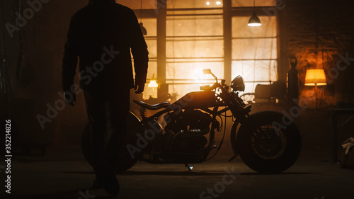 Custom Bobber Motorbike Standing in an Authentic Creative Workshop. Silhouette of a Rider Coming to a Bike. Vintage Style Motorcycle Under Warm Lamp Light in a Garage.