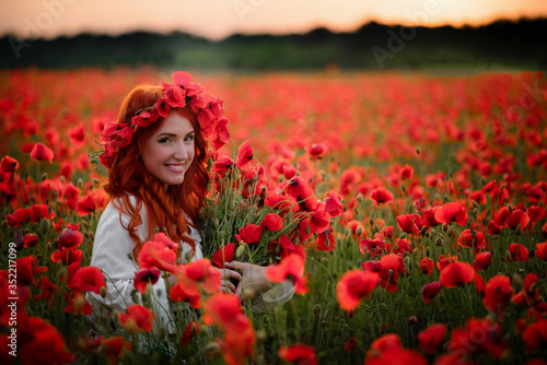 Young female with bouquet of red poppies sitting in flowered field at sunset