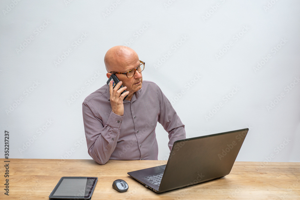 Caucasian male working at home and sitting behind a desk with lap top talking into a cell phone Caucasian male working at home and sitting behind a desk with lap top talking into a cell phone
