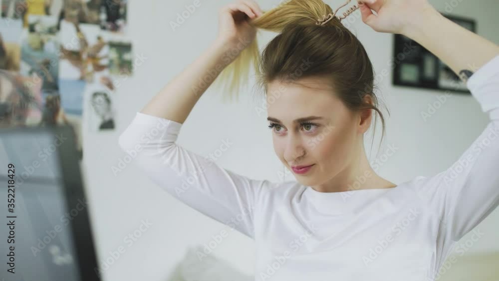 Woman is looking at the mirros and tying her long smooth hair in ponytail, making hairdo