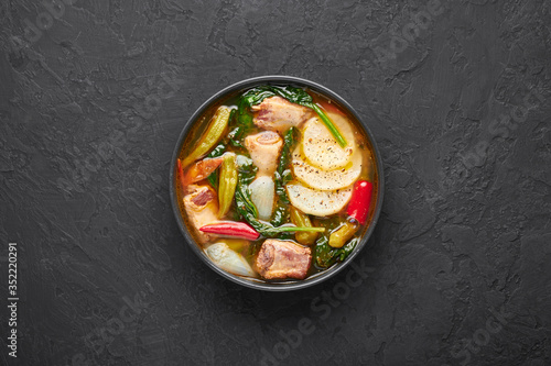 Sinigang na Baboy or Filipino Pork Meat Soup in black bowl on dark slate backdrop. Sinigang is Filipino cuisine dish with meat, bamia, daikon, spinach, fish sauce. Filipino Food. Asian Meal. Top view photo
