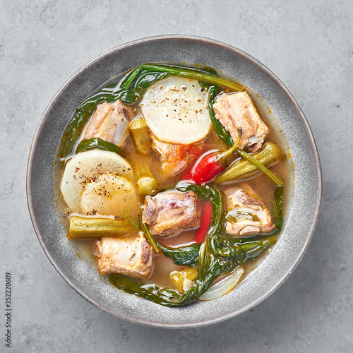 Sinigang na Baboy or Filipino Pork Meat Soup in gray bowl on concrete backdrop. Sinigang is a Filipino cuisine dish with meat, bamia, daikon, spinach, fish sauce. Filipino Food. Asian Meal. Top view photo