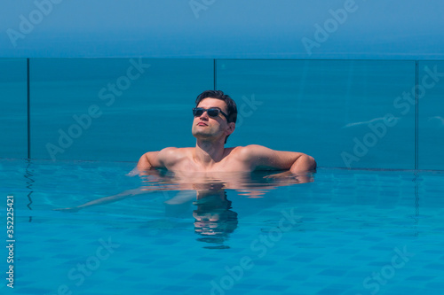 Young caucasian man wearing sunglasses at the edge of an infinity pool with blue sea and sky in the background