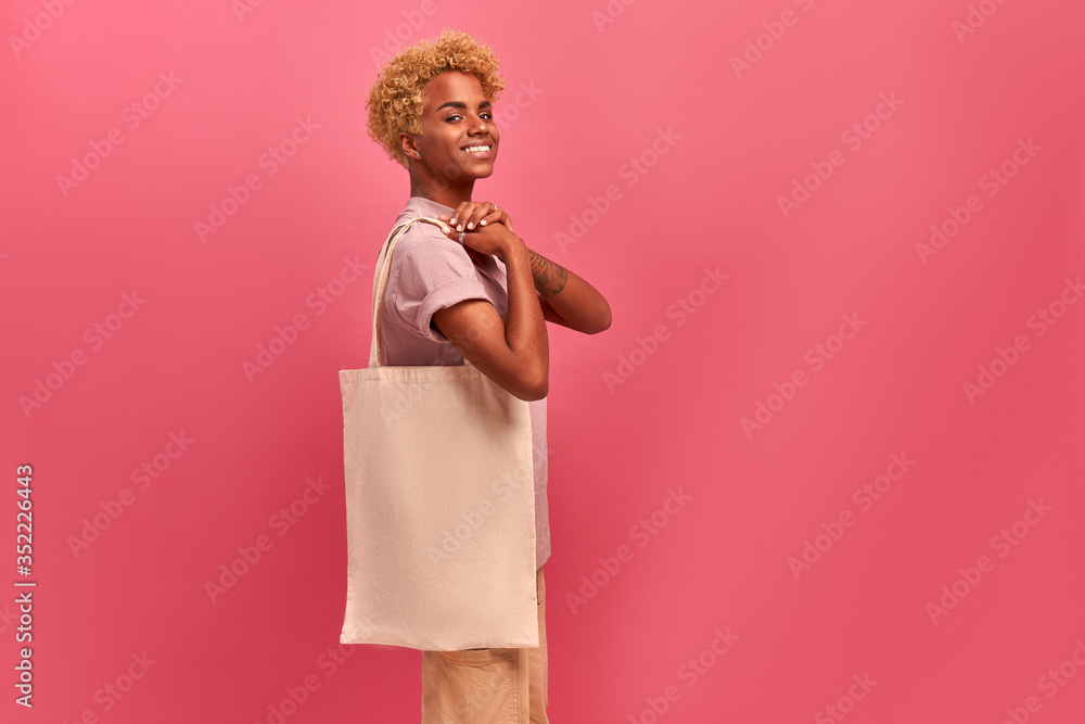 Cheerful smiling african american young woman holding an empty big white woven cotton eco bag in hands for your advertising mockup space on pink background. Green materials concept. Say no to plastic Cheerful smiling african american young woman holding an empty big white woven cotton eco bag in hands for your advertising mockup space on pink background. Green materials concept. Say no to plastic