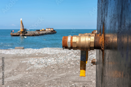 An industrial rusty water faucet on the shore with blurred barge in the sea