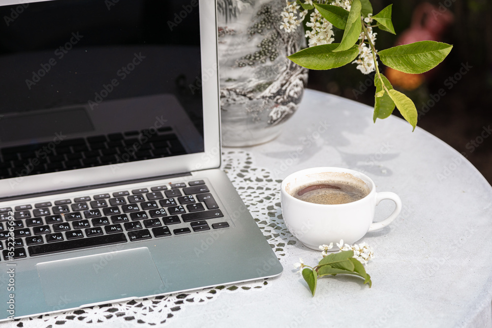 Cup of coffee, laptop, bird cherry color on the table, blurred garden background, green leaves, flowers Cup of coffee, laptop, bird cherry color on the table, blurred garden background, green leaves, flowers