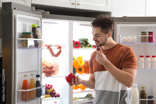 Young man with bell pepper and grape berry near open refrigerator indoors