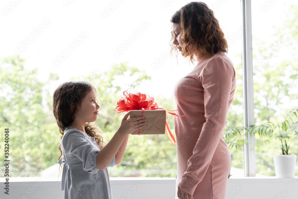 Girl schoolgirl in a blue dress gives her mom a gift. Mom has a birthday. She is smart and happy. The box with a surprise is big.