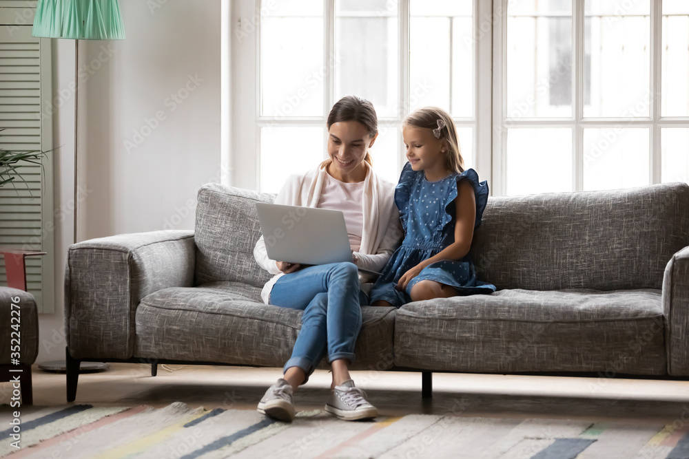 Smiling young mother and little daughter sit relax on sofa in living room using laptop together, happy mom and small girl child rest on couch at home browsing internet shopping on modern computer