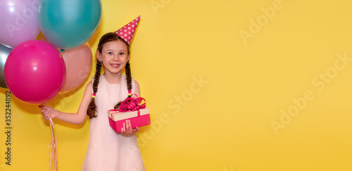 Happy Birthday! Happy girl with balloons and cap holds a gift on a yellow background. Copy space for text. Banner