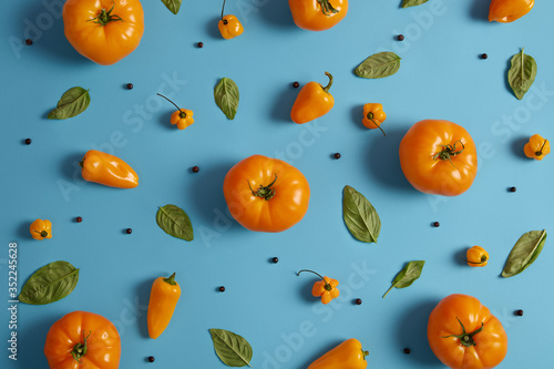Shot of ripe yellow tomatoes, paprika, peppercorns and green leaves of basilic on blue background. Collection of fresh vegetables and spices for cooking vegetarian dish. Natural food concept
