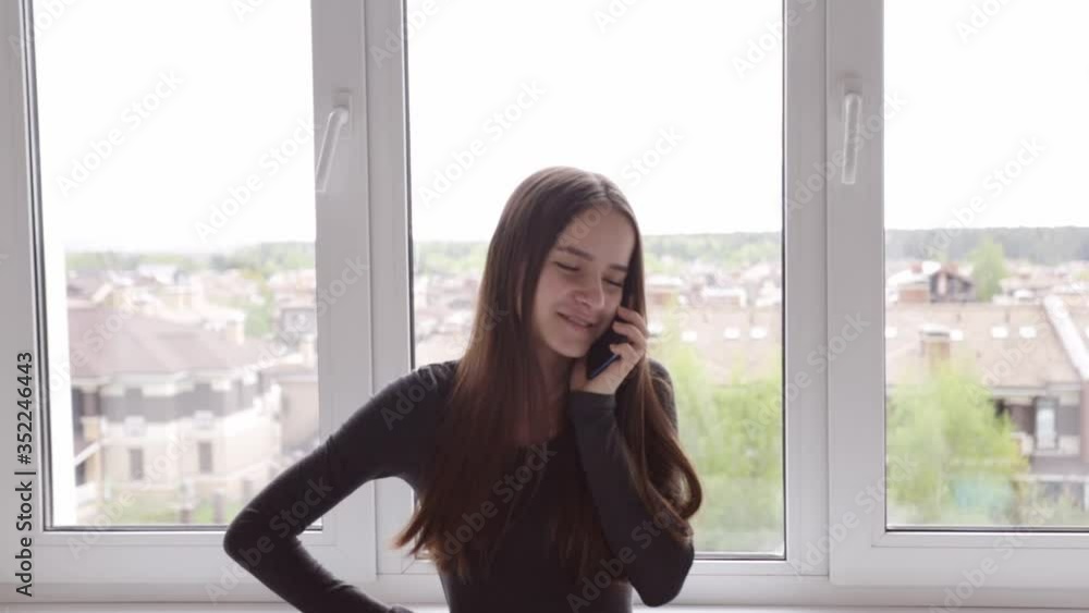 happy, attractive young woman talking on a cell phone at home, standing by the window