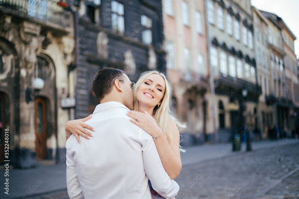Lovely couple walking around the block. Dark-haired man in a white shirt hugging a blonde in a beautiful dress Lovely couple walking around the block. Dark-haired man in a white shirt hugging a blonde in a beautiful dress