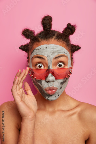 Naked surprised woman applies clay mask for refreshing and reducing wrinkles, wears pink sunglasses, has knot hairstyle, enjoys beauty procedures, poses against rosy background. Skin care, spa