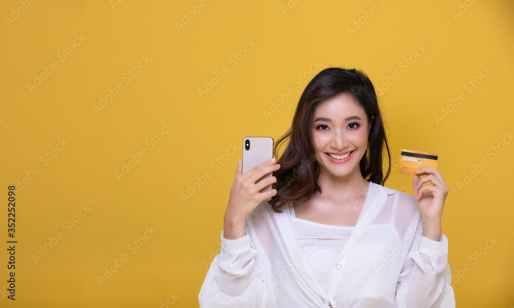 Portrait Asian beautiful happy young woman smiling cheerful and she is holding credit card and using smart phone for shopping online on yellow background. Portrait Asian beautiful happy young woman smiling cheerful and she is holding credit card and using smart phone for shopping online on yellow background.