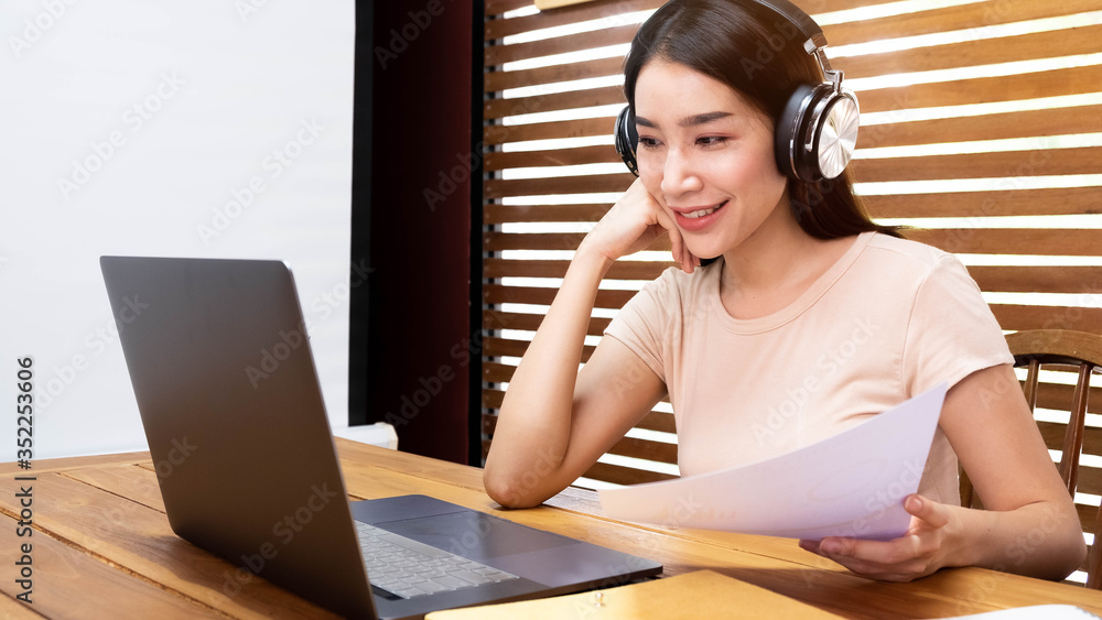 Asian girl in headphones sit at desk watch webinar making notes, happy biracial young woman in earphones work study using computer. Asian girl in headphones sit at desk watch webinar making notes, happy biracial young woman in earphones work study using computer.