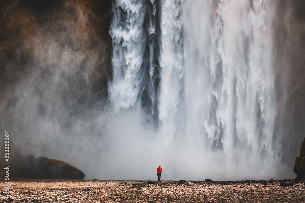 Amazing Icelandic Landscape. Man standing in front of the majestic Skogafoss Waterfall. Incredible view of Skogafoss waterfall during sunset. Dramatic Scenery of Iceland. Best  travel locations