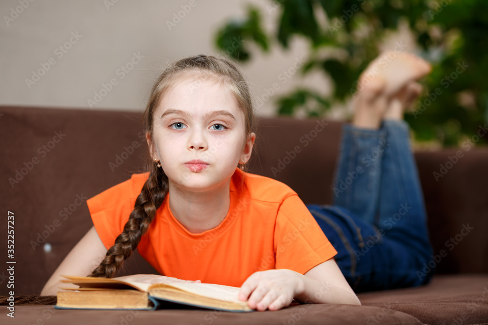 Cute caucasian little girl with a long braid reading book while lying on sofa at home during quarantine due to coronavirus. Education and leisure concept Cute caucasian little girl with a long braid reading book while lying on sofa at home during quarantine due to coronavirus. Education and leisure concept