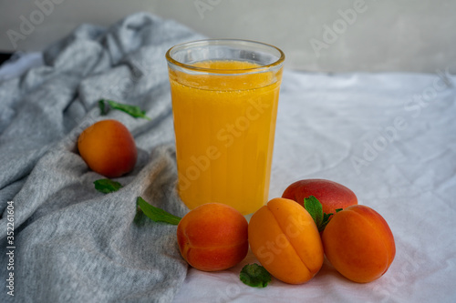 A glass of orange juice surrounded by ripe apricots and mint leaves on textile surface
