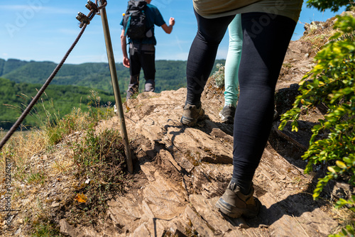 A woman walking a trail through the vineyards on a rocky, slate surface in specialized climbing shoes by a metal railing, in the background stands a man.