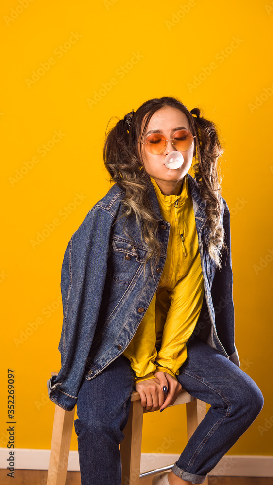 a girl of Asian appearance sits against a yellow wall, inflates large pink bubbles, in a jeans a girl of Asian appearance sits against a yellow wall, inflates large pink bubbles, in a jeans