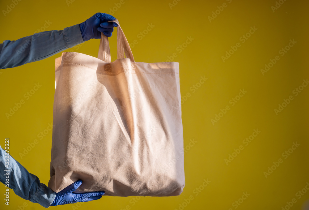 Sending eco-friendly reusable cotton bag on a yellow background. The courier holds a canvas fabric bag without plastic packaging on a yellow background. Safe delivery to the epidemic. Sending eco-friendly reusable cotton bag on a yellow background. The courier holds a canvas fabric bag without plastic packaging on a yellow background. Safe delivery to the epidemic.